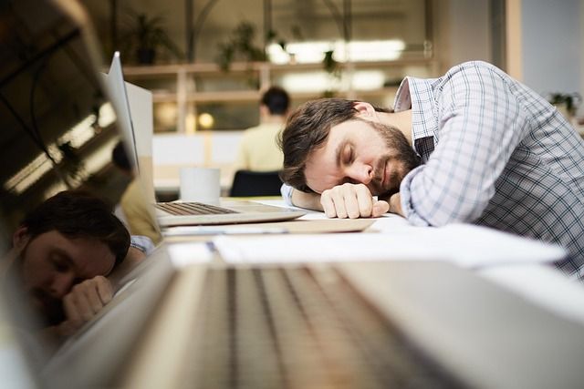 An exhausted office worker leaning on a desk with eyes closed from fatigue.